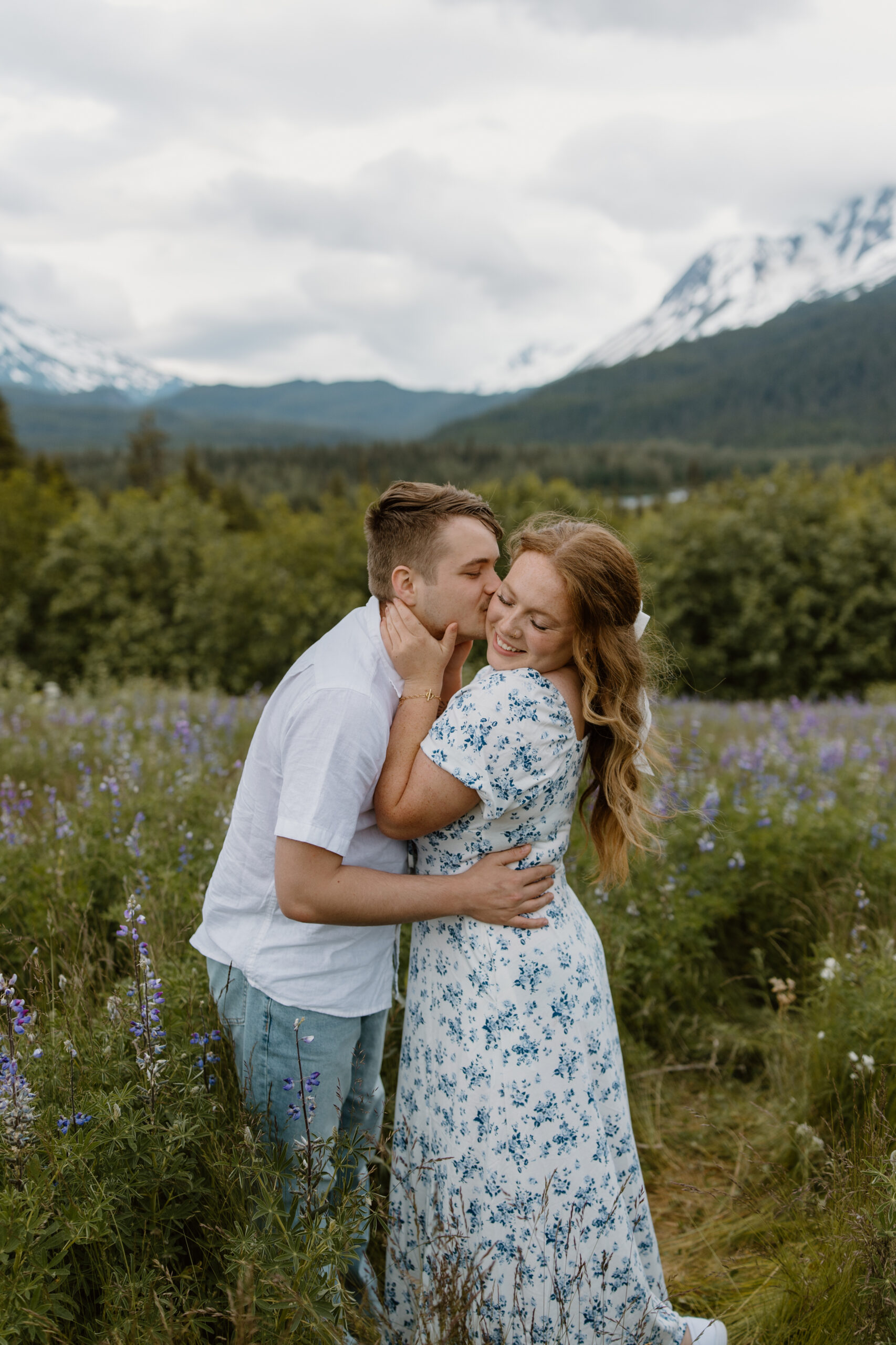 A couple embraces as the boyfriend kisses his girlfriend's cheek. They are standing in a field of lupine with mountains behind them.