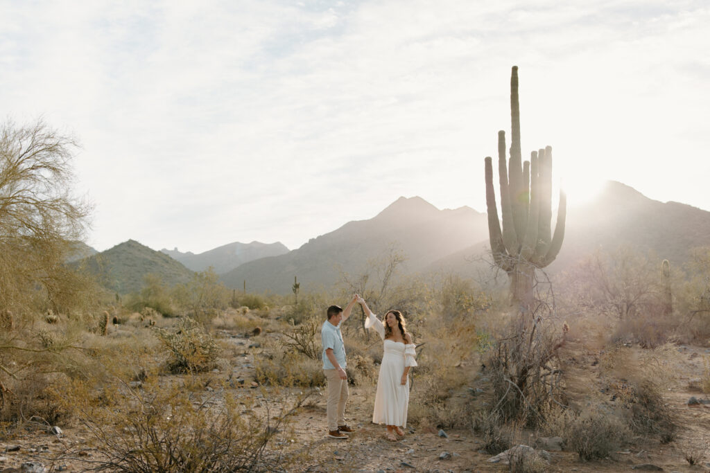 Man spins woman as they smile at each other during a sunrise session at McDowell Sonoran Preserve, Arizona. Mountains in the background and a saguaro cactus beside them.