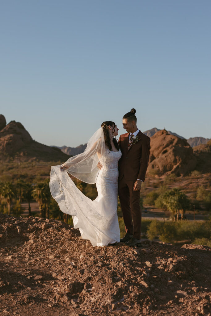 Wedding couple stand next to each other while smiling. Bride is holding the hem of her dress. They are at Papago Park with red rocks behind them.