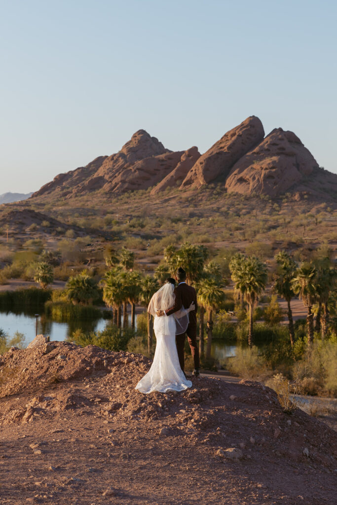 Wedding couple stand next to each with their back towards camera. They are at Papago Park with red rocks in front of them.