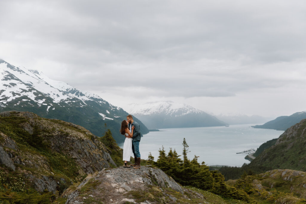 Couple stands on boulder and kiss with Prince William Sound and mountains in the background.