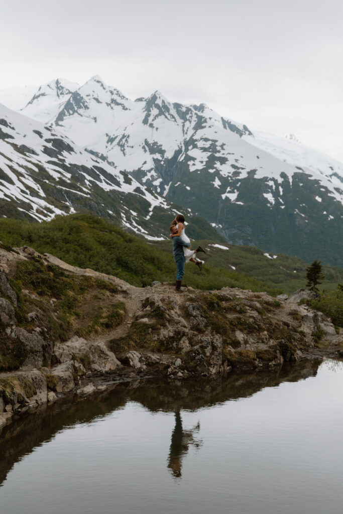 Engaged couple at Portage Pass Alaska, groom picking up bride with an alpine pond in the foreground and mountains in the background.