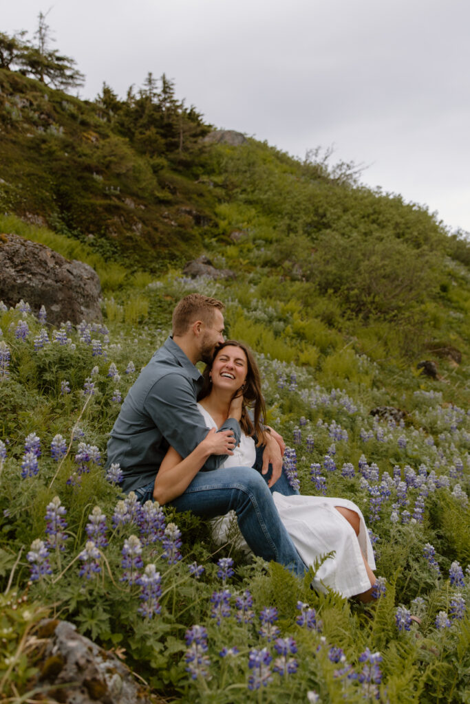 Engaged couple sitting on a hillside covered in wild lupine flowers at Portage Pass, bride leaning into groom's chest as they smile