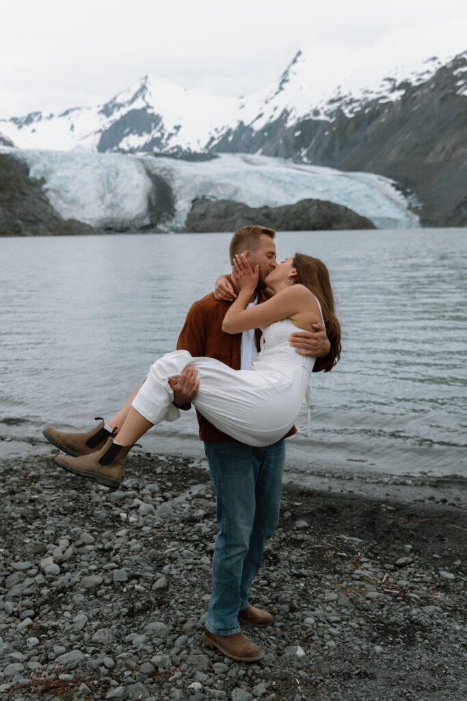 Engaged couple at Portage Lake Alaska, groom picking up bride and kissing her with Portage Glacier in the background
