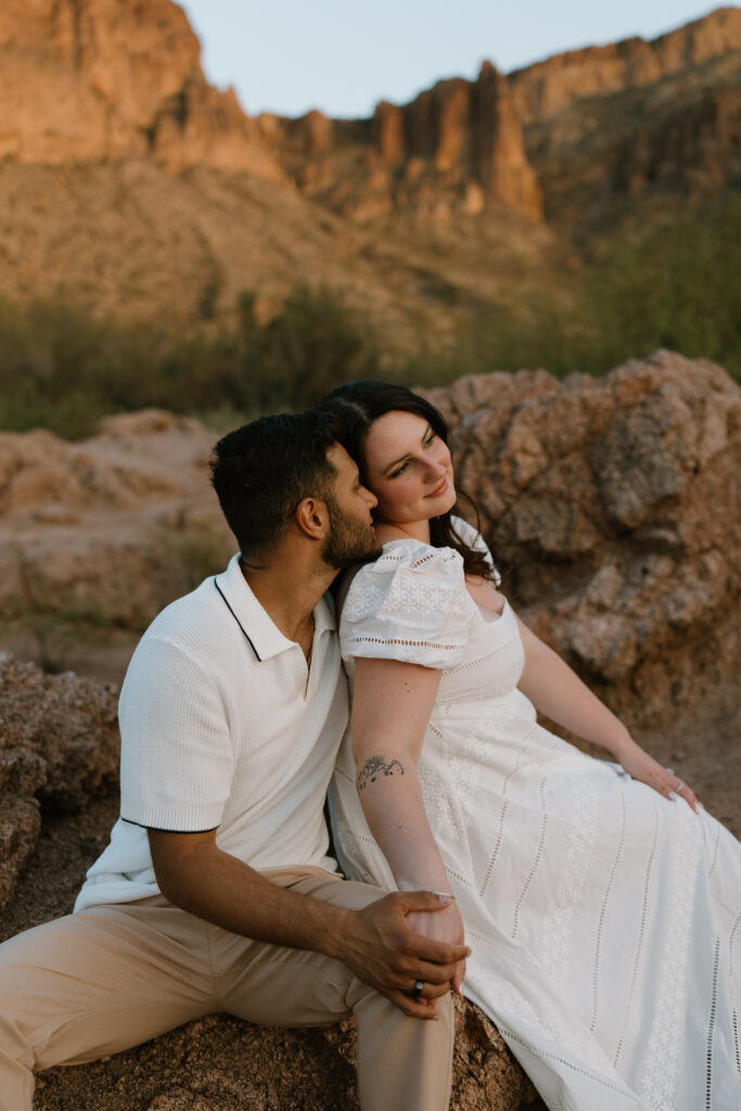 Engaged couple sits on large rocks next to each other while leaning into one another.