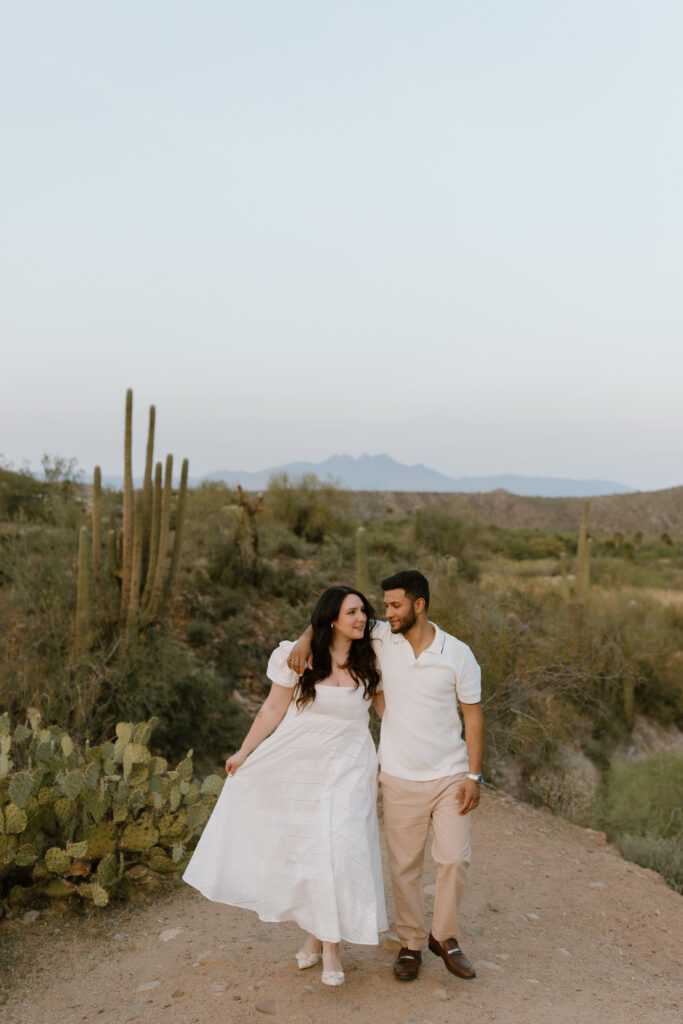 Engaged couple walks together. his arm is around her shoulder and she is holding the hem of her dress. There are cacti behind them.