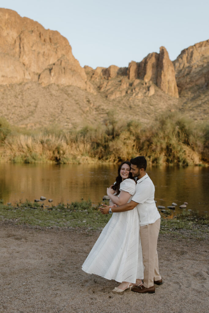 Engaged couple embraces each other near a river. Mountain cliffs are beyond the river.