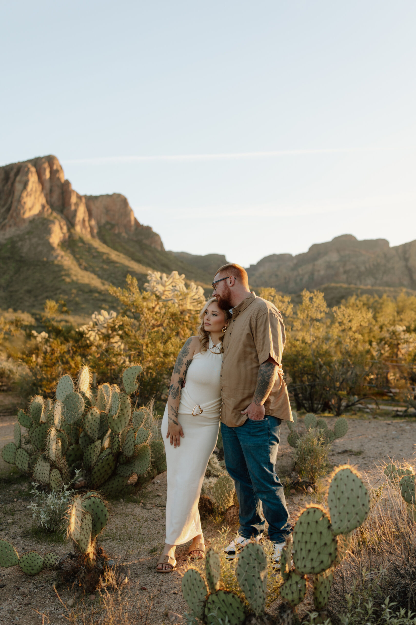 Engaged couple leaning into each other and gazing into the distance at sunset, with Salt River bluffs in the background and prickly pear cacti in the foreground. Phoenix-area engagement session.