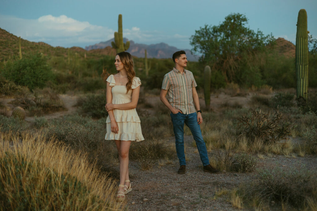 Couple stands near each other looking opposite directions. There are in the desert with saguaros and mountains in the distance.