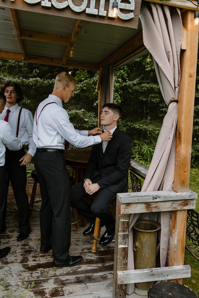 Groom sits on a chair on a porch as his groomsmen adjusts his bowtie.