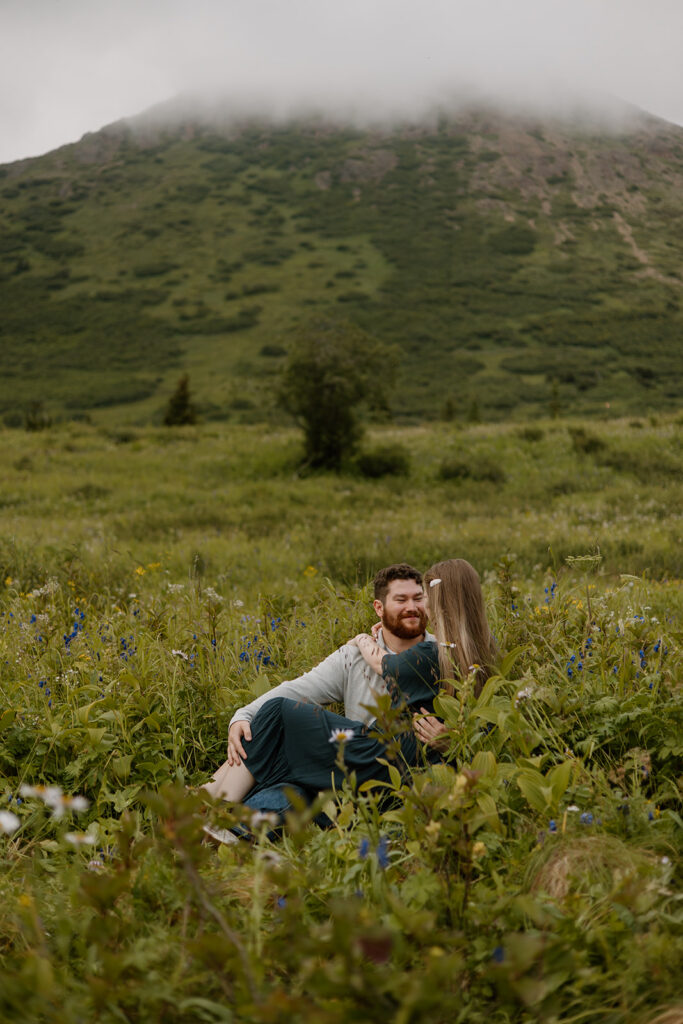 Engaged couple laughing together in a green mountain valley in South Central Alaska, bride sitting in groom's lap as they smile at each other.