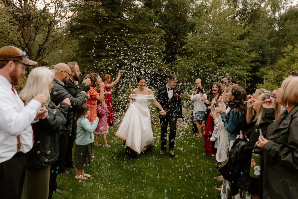 Bride and groom run through confetti and bubbles thrown by their guests as they exit their wedding. They are standing on a grass lawn with pine trees behind them.