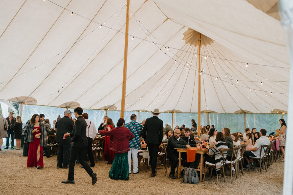 Guests are sitting and standing in the reception space that is covered by a canopy tent.