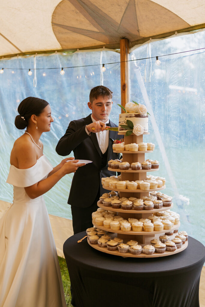 Groom cuts into wedding cake while the bride holds a plate. There is cupcakes stacked as a tower beneath the cake.