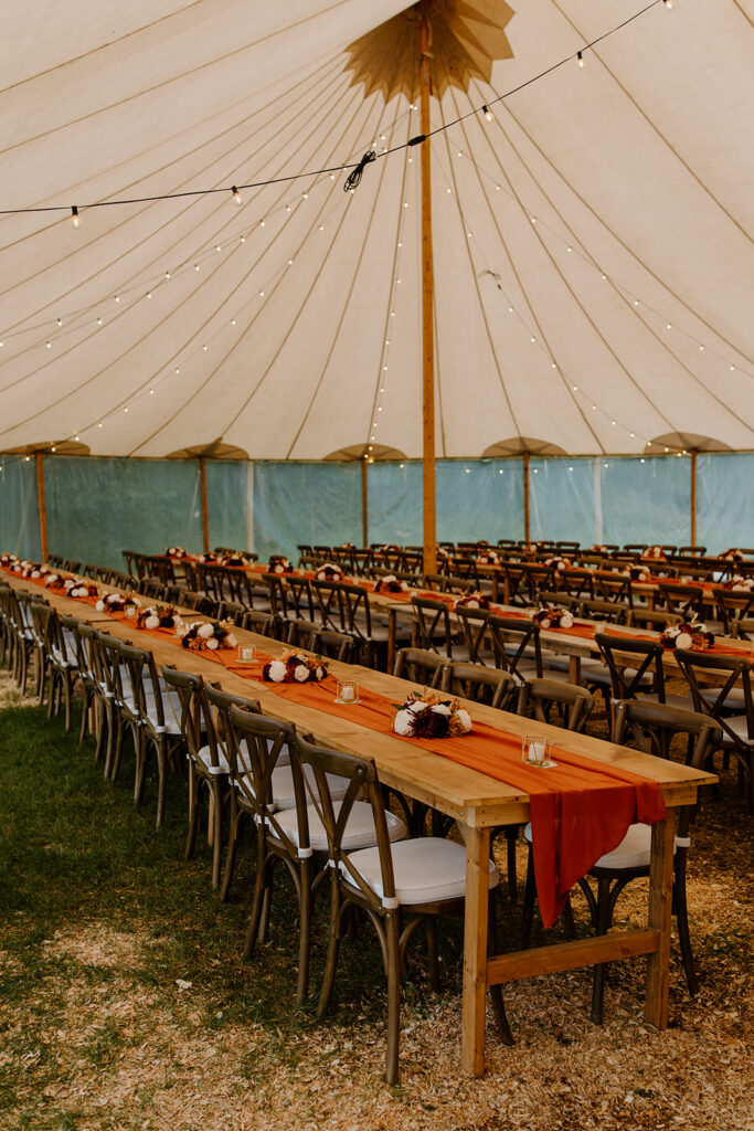 Inside of a wedding canopy tent with strung lights on the ceiling. Several rows of feast tables and chairs are decorated with fall colored table settings.