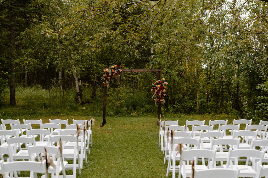 Wedding arch with flowers on the sides at the front of the alter. White chairs line the aisle leading to the arch. Green forest and grass surrounding.