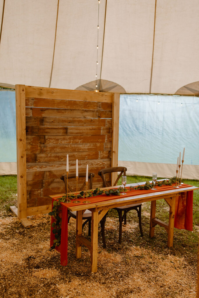 The sweet heart table for the bride and groom decorated with candles, an orange table runner, and greenery. A wood backdrop is behind the table.