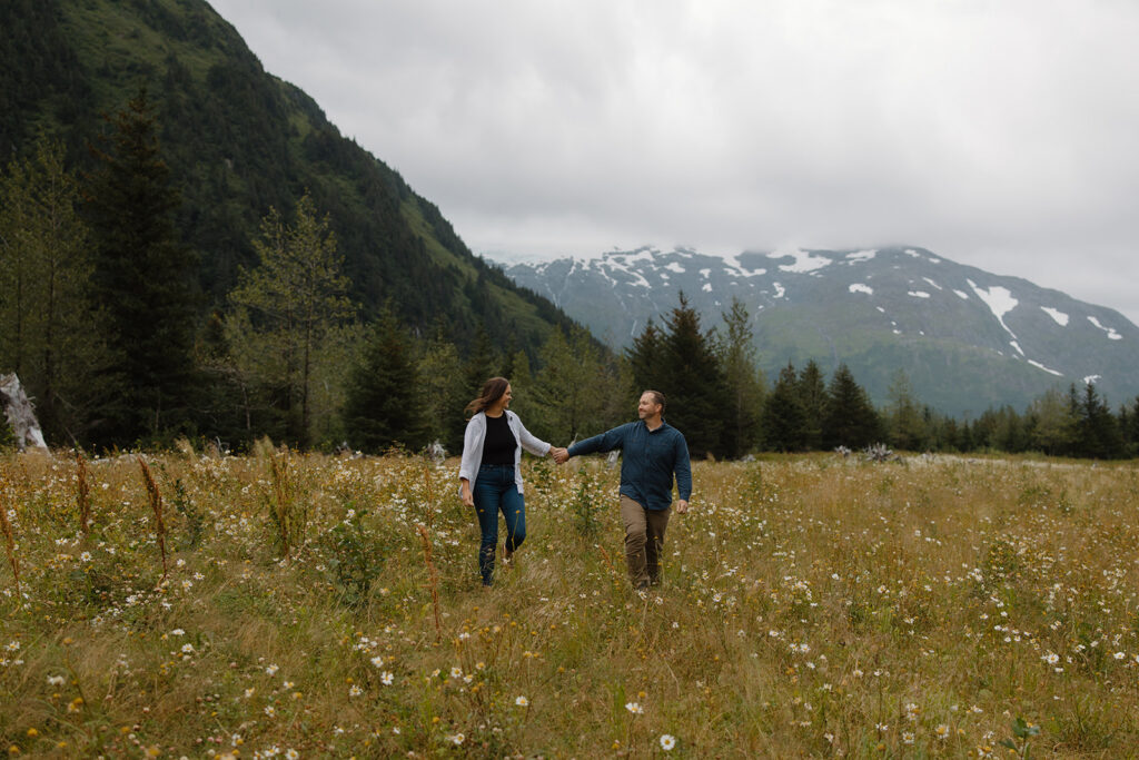 Married couple walks holding hands at Portage Alaska, bride in groom's lap smiling at each other with daisies on the hillside on an overcast day