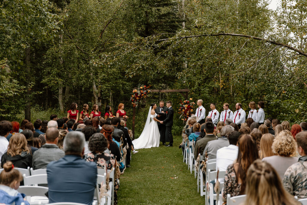 Bride and groom are holding hands at the alter surrounded by a forest. The guests are in sitting in chairs watching.
