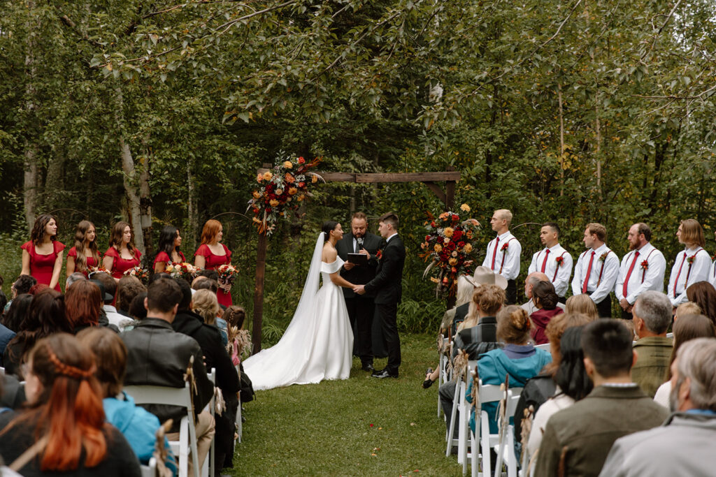 Bride and groom are holding hands at the alter surrounded by a forest. The guests are in sitting in chairs watching.