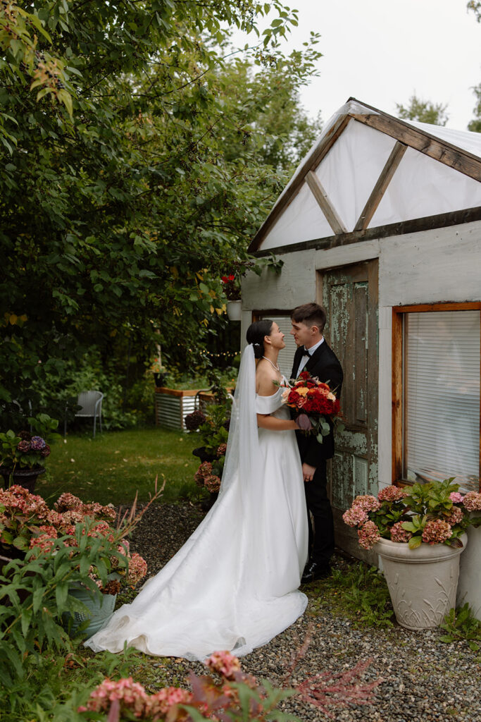 Bride and groom stand close to one another smiling at each other. They are standing next to a green house and surrounded by green trees and pink flowers.