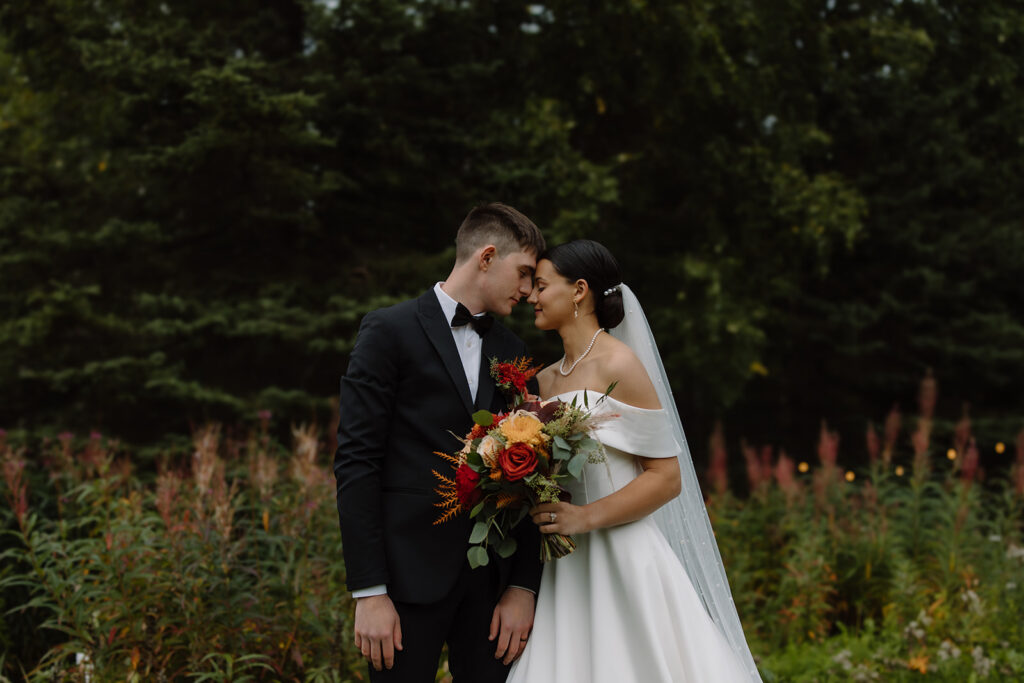 Bride and groom touch foreheads together with their eyes closed, surrounded by trees and fireweed. 