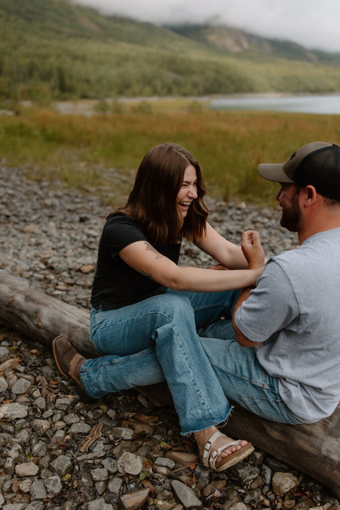 Engaged couple straddling a log on the rocky beach at Eklutna Lake Alaska, bride playfully shoving groom as they smile