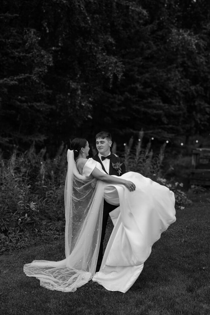Black and white photo of groom picking up bride. Her veil is drapped behind her.