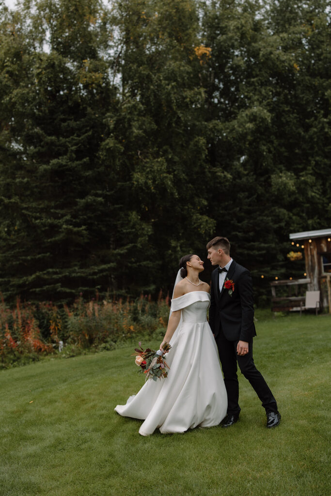 Bride and groom lean in towards each other for a kiss. They are standing on a grass lawn with pine trees behind them.