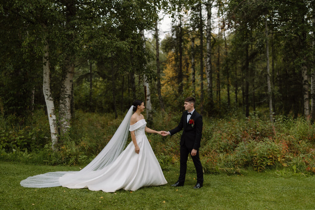 Bride and groom reaches hands towards each other while looking at one another. They are standing on a grass lawn with birch trees behind them.