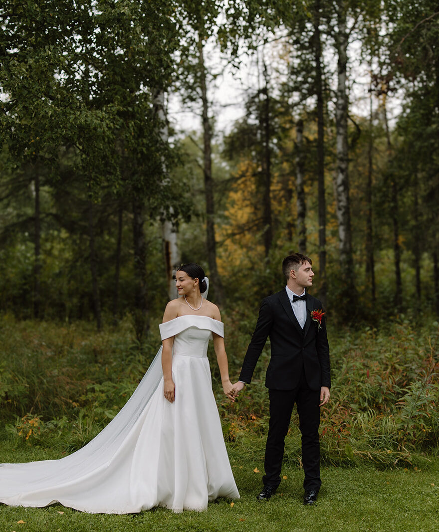 Bride and groom holds hands while looking out opposite directions. There is a green forest behind them and they are standing on grass