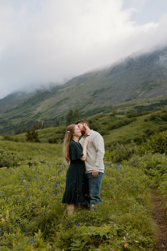 Engaged couple embracing in a green mountain valley in South Central Alaska, groom kissing bride on the cheek as she smiles