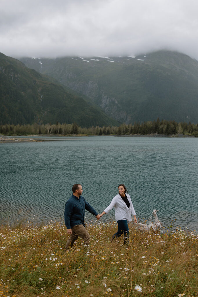 Married couple walks along the shoreline holding hands at Portage Alaska, bride in groom's lap smiling at each other with daisies on the hillside on an overcast day