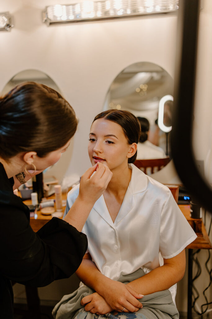 Makeup artist puts lipstick on bride as she sits in her silk pajamas.