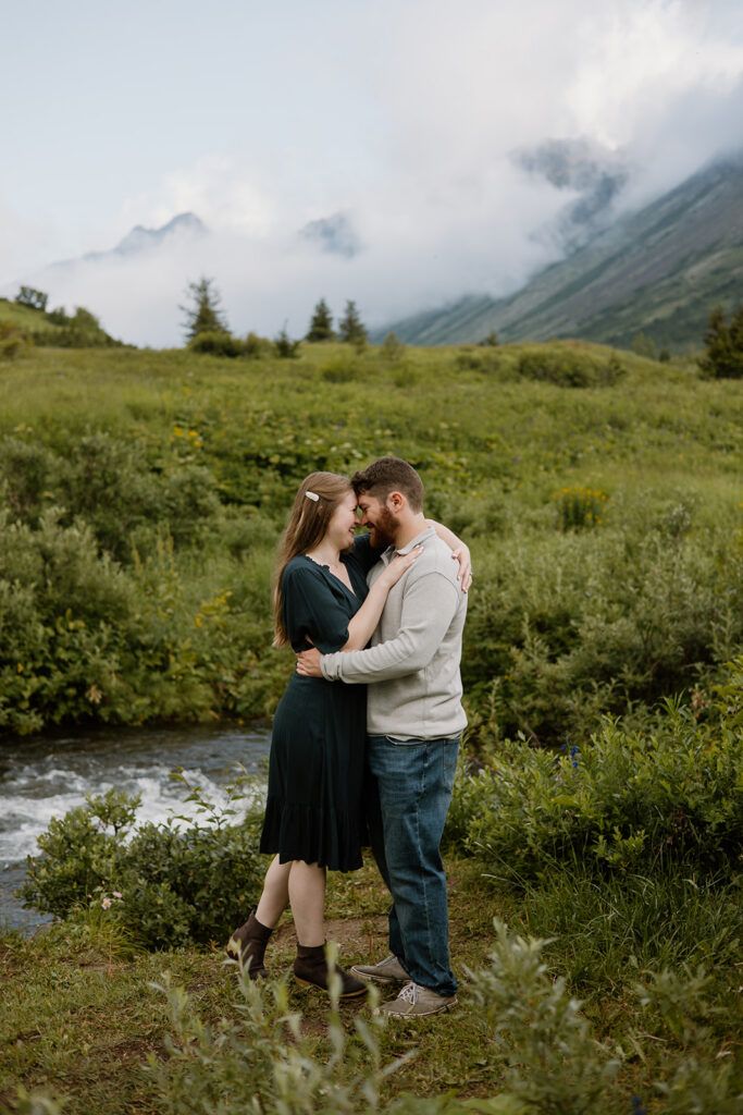 Engaged couple embracing in a green mountain valley in South Central Alaska, couple put foreheads together as they smile.