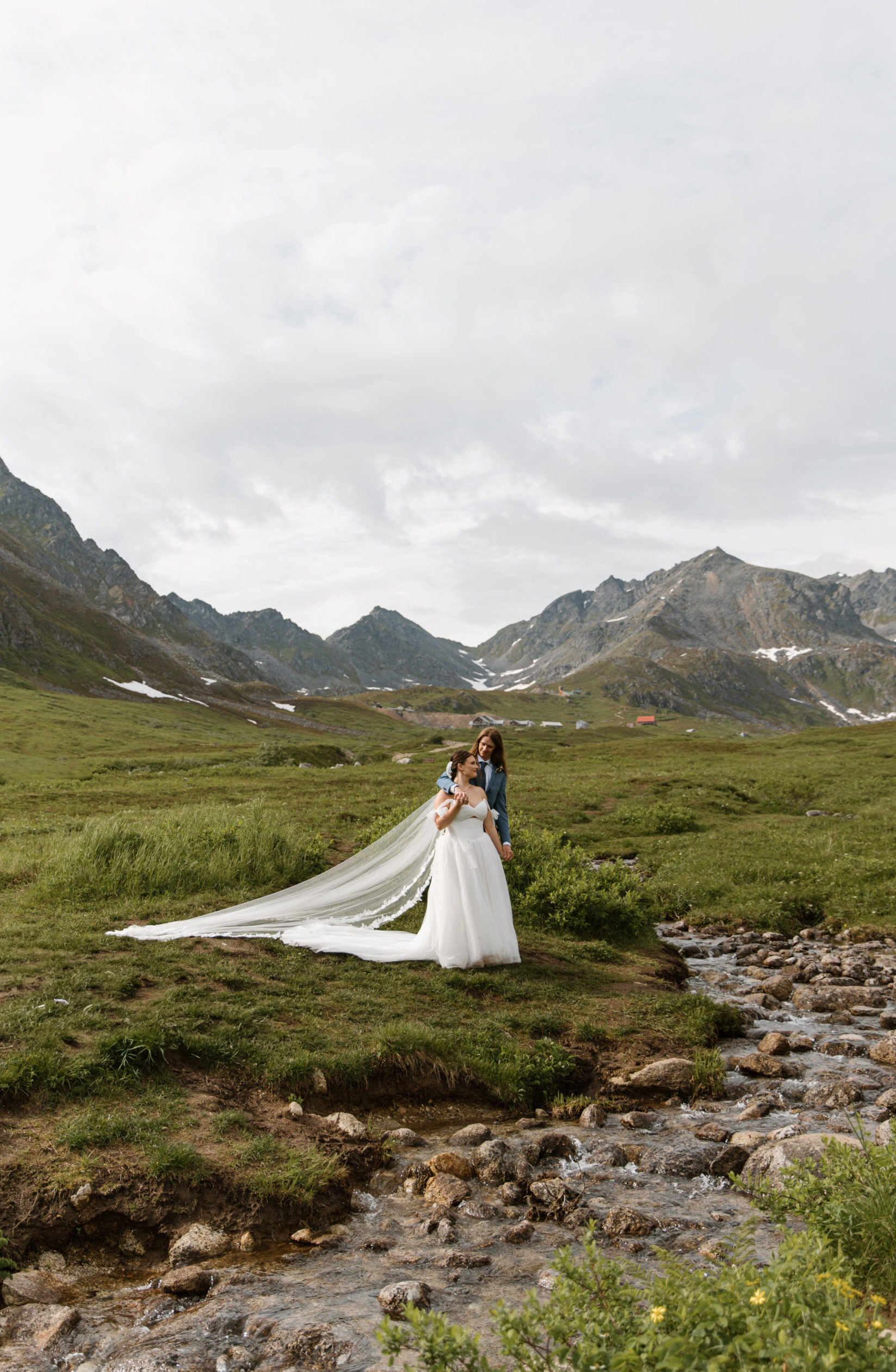 Bride and groom during their Hatcher Pass elopement, groom wrapping around bride and giving her a forehead kiss beside a stream with jagged mountains behind them, veil draping in the breeze
