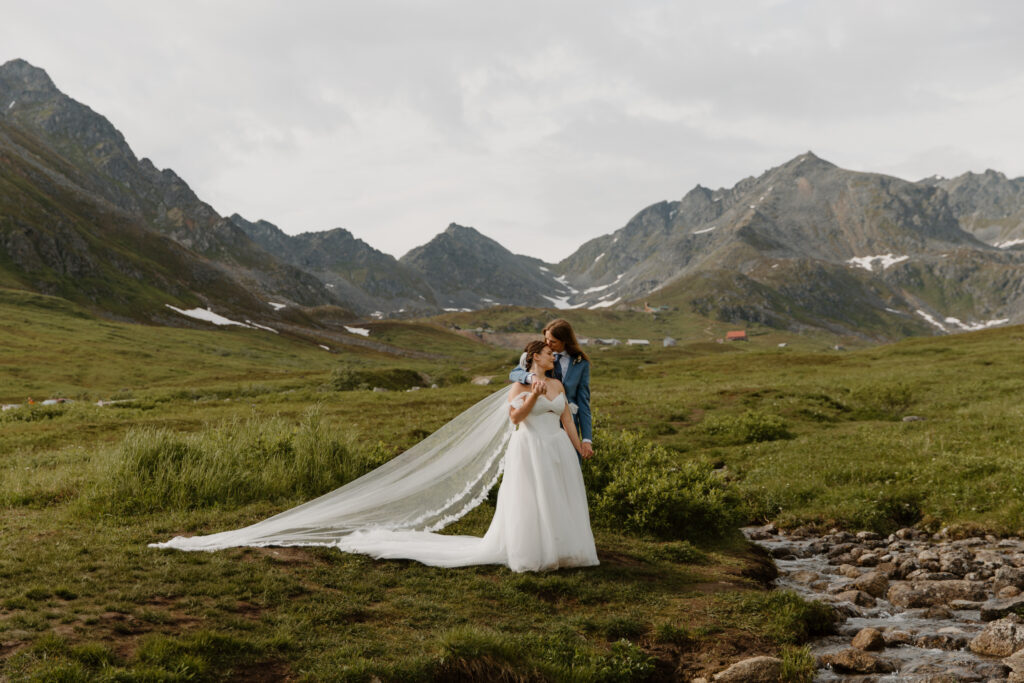 Bride and groom during their Hatcher Pass elopement, groom wrapping around bride and giving her a forehead kiss beside a stream with jagged mountains behind them, veil draping in the breeze