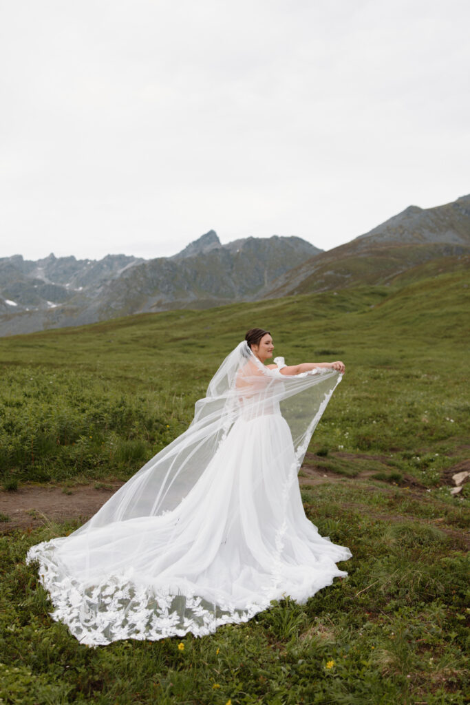 Bride holding up her wedding veil as it cascades down her side in the alpine bowl during her Hatcher Pass elopement in Alaska