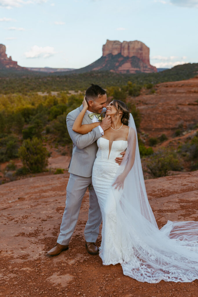 Bride and groom smiling at Cathedral Rock trail in Sedona Arizona, groom standing behind bride holding her chin as she reaches back to touch his face