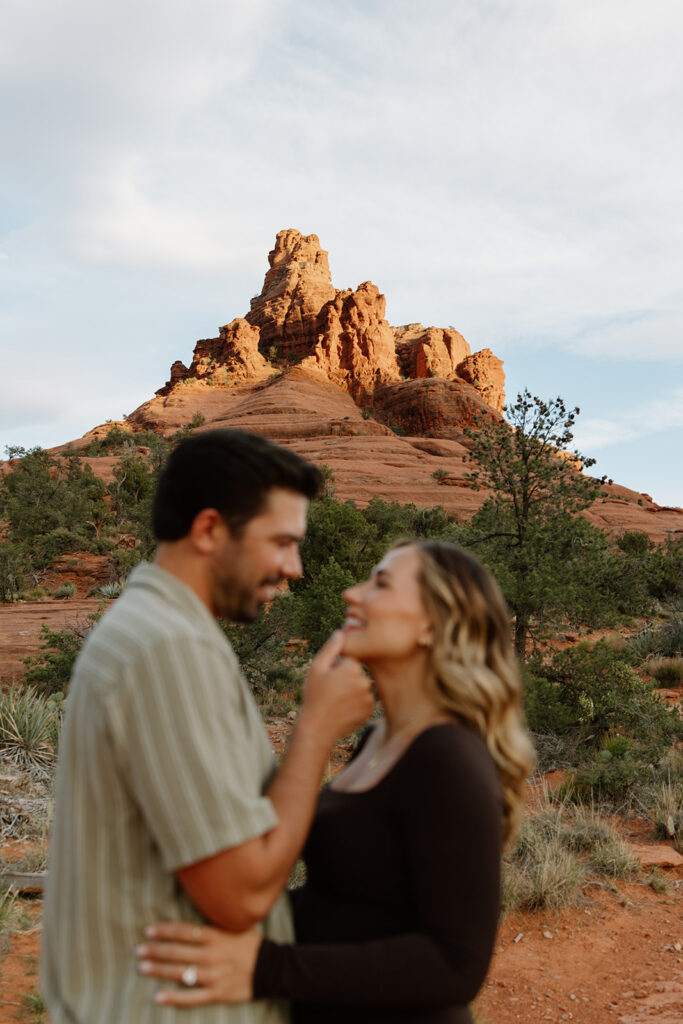Couple smiling as he holds her chin with Bell Rock formation in the background on the Bell Rock trail in Sedona Arizona