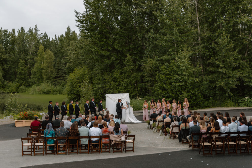 Wide shot of an outdoor wedding ceremony at the Alaska Native Heritage Center in Anchorage Alaska with guests gathered in front of the lake