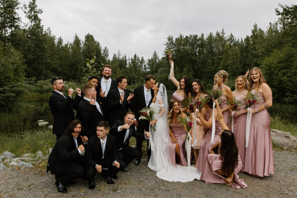 Wedding party cheering as bride and groom kiss with the lake behind them at the Alaska Native Heritage Center wedding in Anchorage Alaska
