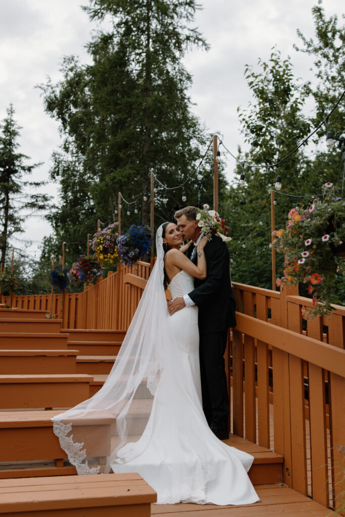 Groom kissing bride on the cheek as they stand on the steps surrounded by hanging flowers during their Alaska Native Heritage Center wedding in Anchorage Alaska