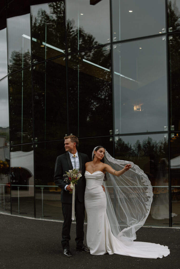 Bride fanning her lace veil in front of the glass museum windows during wedding portraits at the Alaska Native Heritage Center in Anchorage Alaska