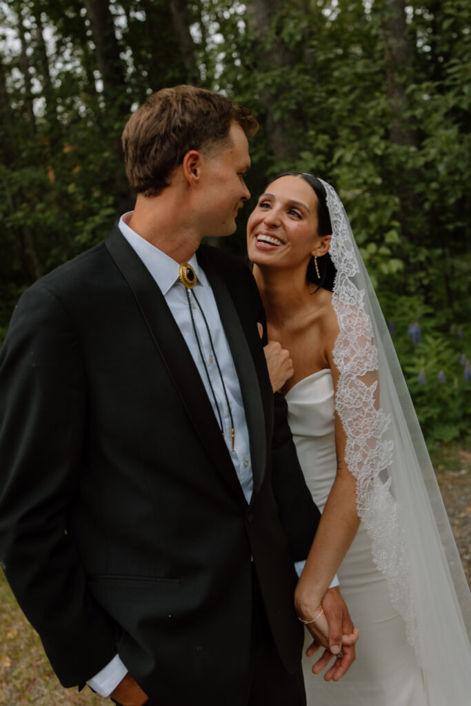 Bride and groom smiling at each other during wedding portraits with a forest backdrop