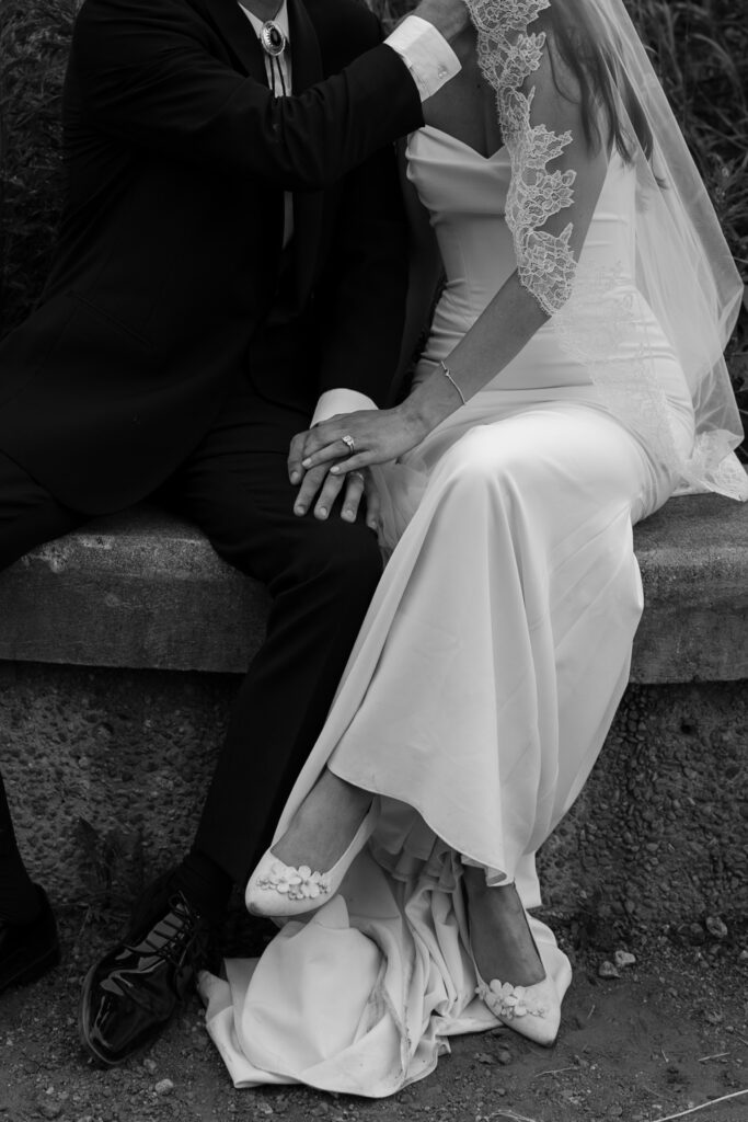 Black and white close up of bride and groom holding hands and leaning in for a kiss while sitting on a bench