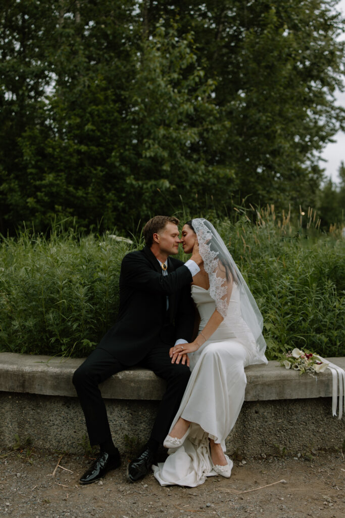Bride and groom holding hands and leaning in for a kiss while sitting on a bench