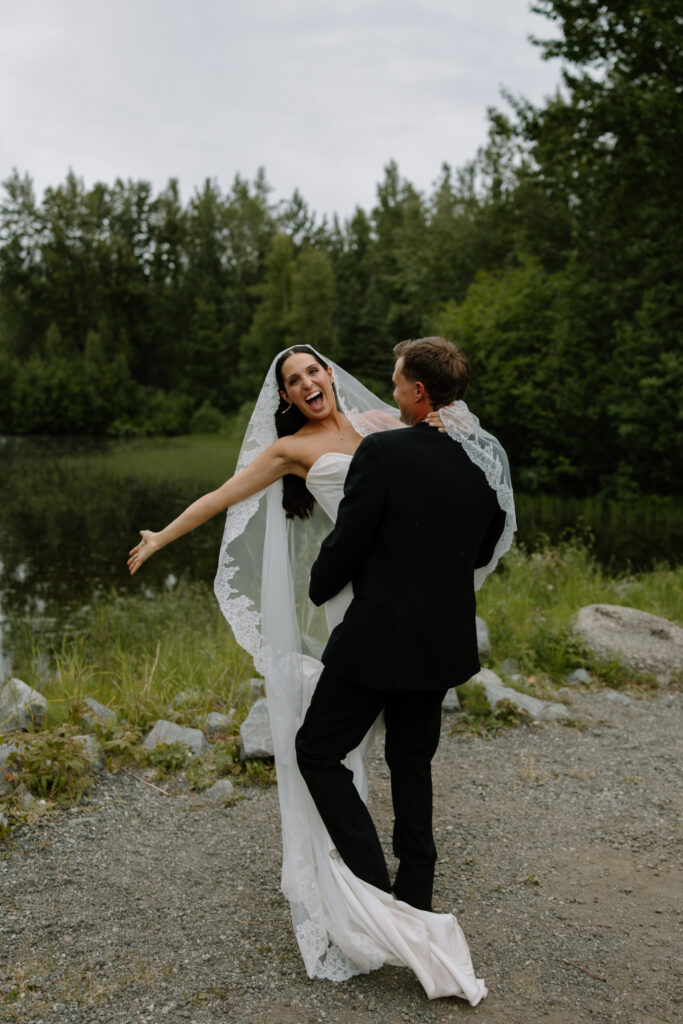 Groom twirling bride as she smiles and waves her arm in the air with a lake and forest behind them