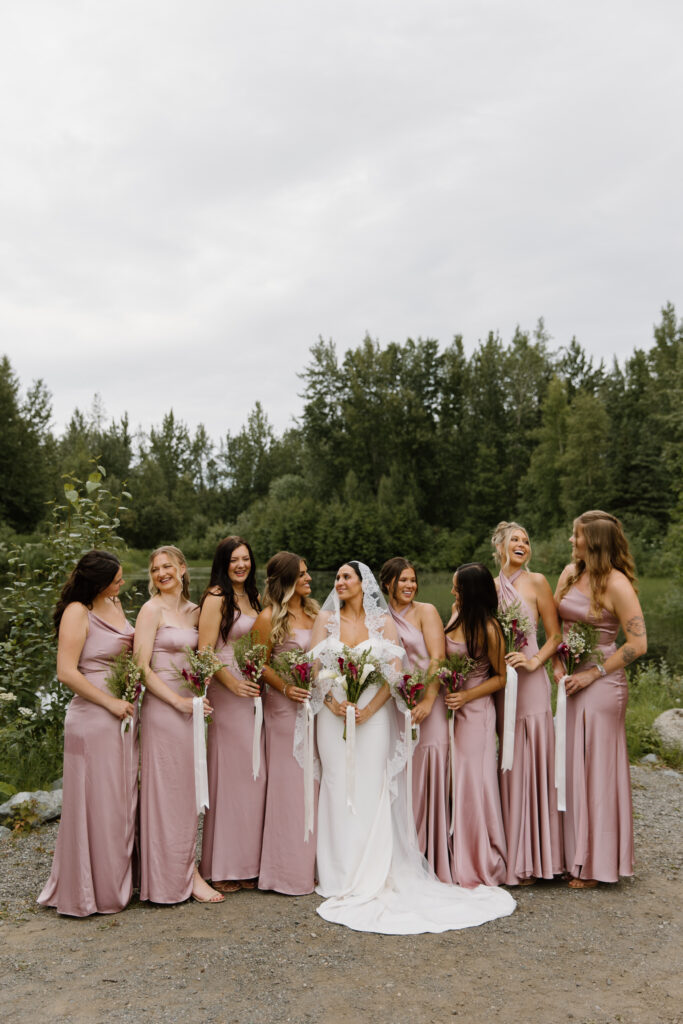 Bride surrounded by bridesmaids in english rose silky dresses smiling at each other during wedding portraits at the Alaska Native Heritage Center in Anchorage Alaska